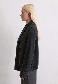 Strickkarte in Dunkelgrau mit einem Schalkragen, ausgestattet mit einem strukturierten Muster und lockerem Schnitt. Das Modell trägt ein passendes graues Outfit.