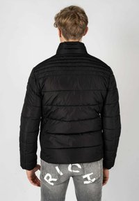 Giacca a vento nera con design a rombi orizzontali, colletto alto e una texture liscia. La vista posteriore mostra una forma aderente e linee pulite.