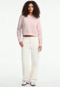 Sweatshirt cropped rosa com texto branco, combinado com calças cargo largas em creme com bolsos laterais e uma textura suave. Ténis brancos.