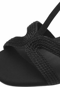Svart sandal med flätade svarta remmar och platt sula. Texturen är slät, med mjuka repdetaljer som korsar varandra i mitten.