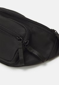 Sac banane en tissu noir avec deux compartiments zippés et languettes sur fond blanc.