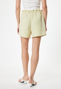 Lys gule, højtsiddende shorts med en flæse i taljen, lavet af glat stof. Modellen bærer hvide hæle sandaler.