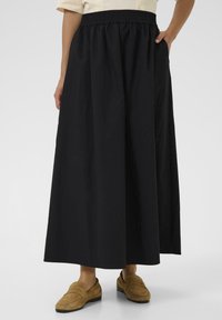 Egy hosszú fekete szoknyát viselő n�ő, rugalmas derékgumival, barna szuéd mokaszinban áll, az egyik kezét zsebébe dugva.