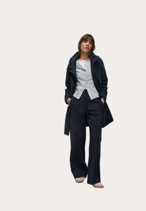 Lagdelte outfit bestående af en mørk hættejakke, grå cardigan og brede sorte bukser. Bløde materialer og afslappet pasform med knapdetaljer.