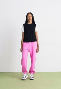 Svart ermeløs topp over en hvit t-skjorte, kombinert med rosa joggebukser og svart-hvite sneakers, mot en ensfarget bakgrunn.