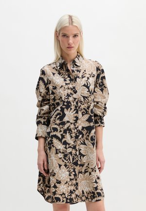 Frau mit glatten blonden Haaren, die ein langärmliges schwarzes Kleid mit beigen Blumen- und Blattmustern trägt und vor einem schlichten weißen Hintergrund steht.