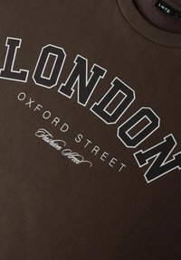 Brun sweatshirt med stor hvid tekst "LONDON" og mindre tekst "OXFORD STREET" i en fed skrifttype, med en script-accent nedenunder.