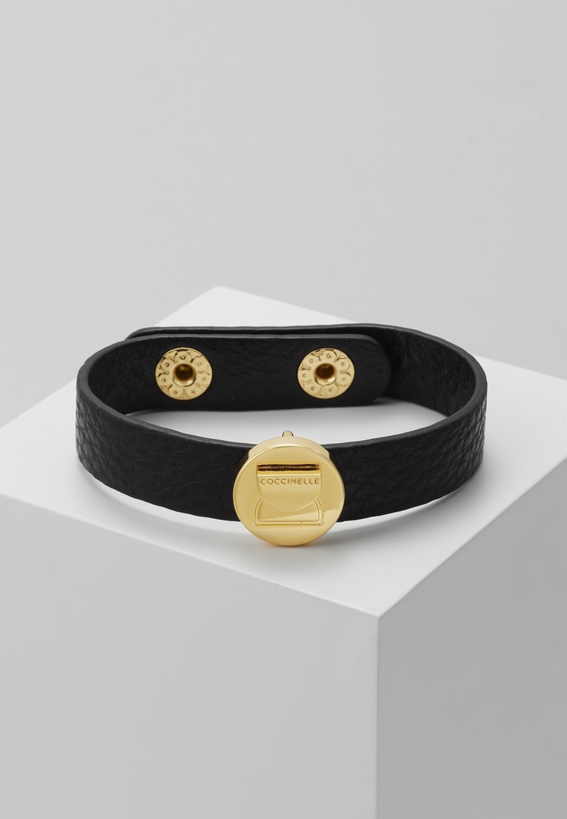 Bracelet en cuir noir texturé avec fermoir circulaire doré gravé "COCCINELLE" et deux boutons-pression dorés pour la fermeture.