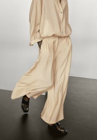 Lösa, ljusbeige byxor med texturerad yta, kombinerade med en matchande skjorta med knappar. Svarta sandaler har en glänsande detalj på remmen.