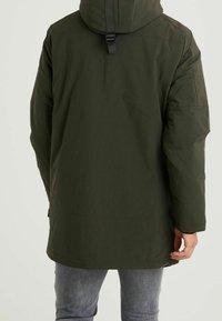 Parkas w kolorze ciemnej zieleni o średniej długości z kapturem, charakteryzujący się gładką, matową teksturą i prostym designem. Posiada tylny pasek do regulacji.