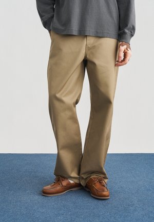 Καφέ χακί βαμβακερό παντελόνι με ίσια γραμμή και ελάχιστες λεπτομέρειες, σε συνδυασμό με καφέ δερμάτινα παπούτσια boat με λευκά κορδόνια και σόλες.
