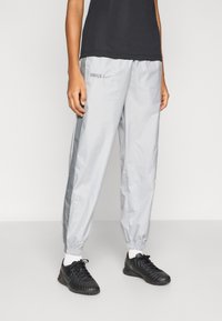 Šviesiai pilkos vėjo nepraleidžiančios kelnės su elastinėmis rankogaliais, tamsiai pilkomis šoninėmis juostelėmis ir aptemptu siluetu. Dėvimos su juodais sportiniais batais ir juodu viršutiniu drabužiu.