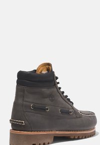 Bottine montante en cuir gris avec semelle marron, bout rond et accents noirs. Présente un design à lacets et un logo visible sur le côté.