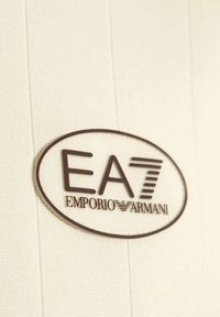 Tessuto bianco con trama verticale presenta un logo ovale prominente in marrone, che mostra "EA7 EMPORIO ARMANI" in caratteri stilizzati.