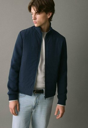 Hombre joven con chaqueta azul marino con cremallera, suéter gris claro y jeans azul claro, mirando hacia su izquierda contra un fondo gris liso.