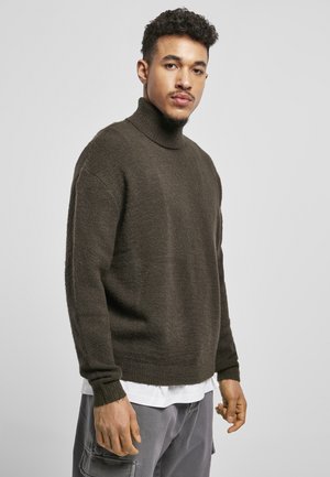 Suéter de cuello alto de punto marrón oscuro, con un corte holgado y puños acanalados, llevado sobre una camisa blanca, con una tela texturizada.