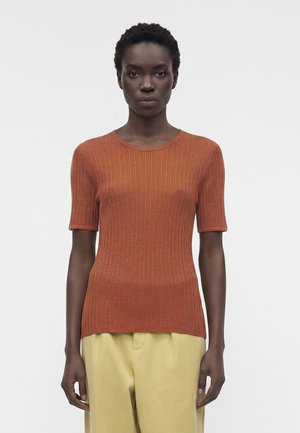 Γυναίκα με κοντά σγουρά μαλλιά που φοράει πουλόβερ κοντομάνικο σε σκουριά απόχρωση με ραβδώσεις και ανοιχτό μπεζ παντελόνι, στέκεται μπροστά σε απλό φόντο.