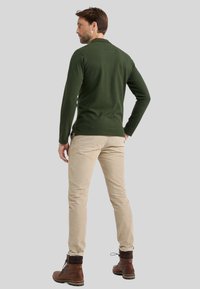 Donkergroene lange mouw polo met zijsplitten, gecombineerd met lichtbeige broeken en bruine veterschoenen, gezien van achter.
