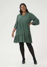 Vestido verde texturizado con escote en V, mangas tres cuartos y dobladillo en capas, llevado sobre mallas negras y tacones de punta.