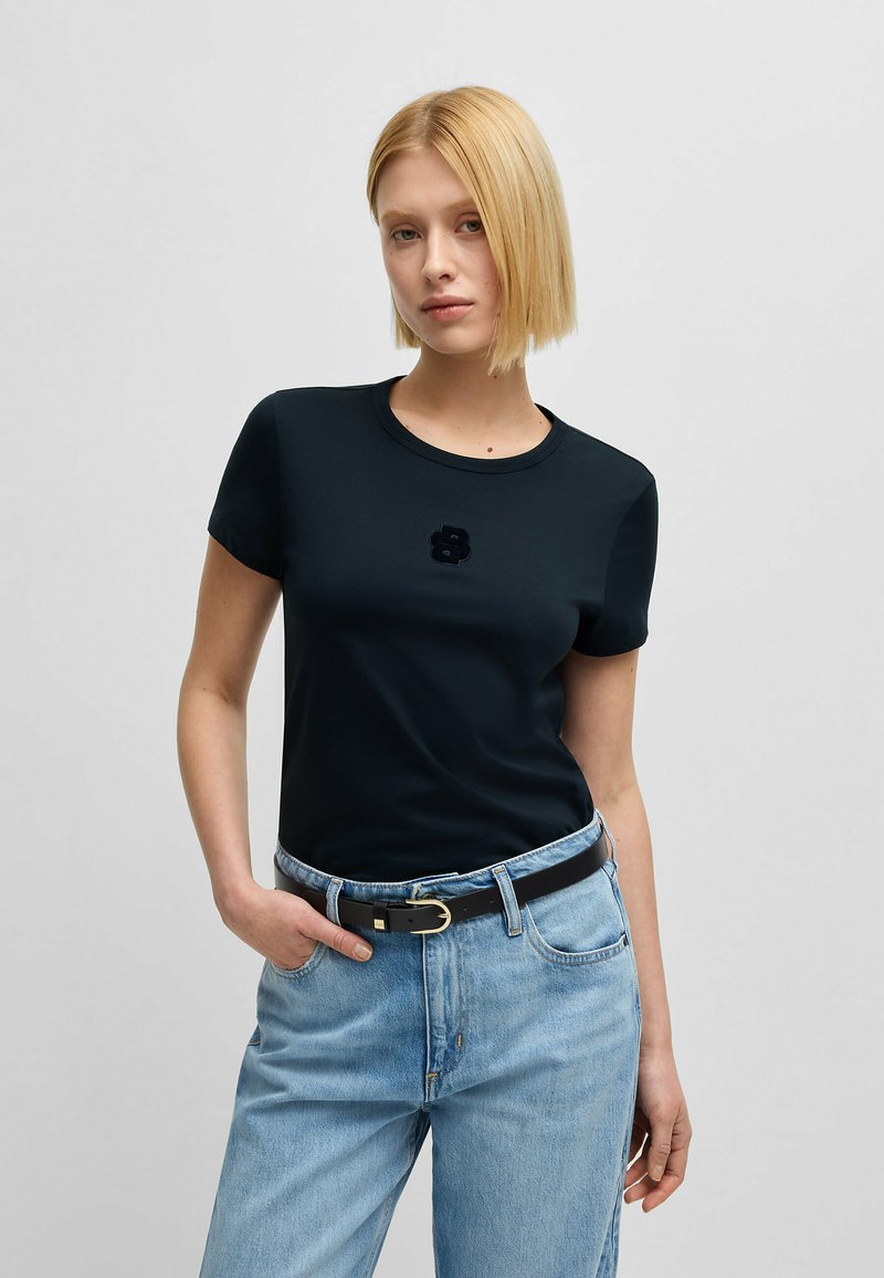 Czarny t-shirt z krótkim rękawem i okrągłym dekoltem, ozdobiony teksturowym emblematem numeru 8 na piersiach, zestawiony z jasnoniebieskimi dżinsami i czarnym paskiem.