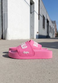 Paire de sandales slides Buffalo roses avec logo blanc posées sur un trottoir en béton devant un bâtiment en briques blanches.