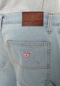 A világoskék farmernadrág barna bőr címkével rendelkezik, melyen a "GUESS JEANS" felirat olvasható, a hátsó zsebén piros háromszög alakú logó található, valamint narancssárga varrás díszíti.