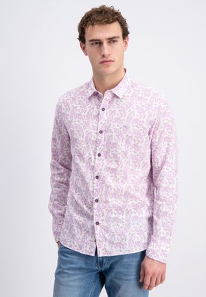 Młody mężczyzna w białej koszuli z długim rękawem z różowym wzorem liści i niebieskich dżinsach, stojący z rękami w kieszeniach, patrzący na bok.