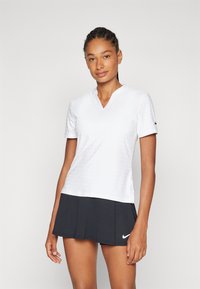 Balta trumpa V formos iškirptės marškinėliai su subtiliu taškiniu raštu, derinami su juoda sijonu. Ant rankovės matomas Nike logotipas.
