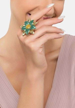 Gold floral ring featuring green and yellow accents with textured petals. Hand model displays ring against a soft pink background.