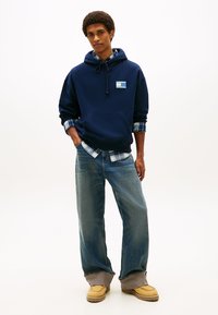 Μπλε φούτερ με κουκούλα, με μπροστινή τσέπη και τετράγωνο έμβλημα, φορεμένο πάνω από μπλε καρό πουκάμισο φανέλα, σε συνδυασμό με άνετο ανοιχτό μπλε τζιν και μπεζ παπούτσια.