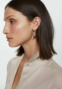 Woman with straight, dark hair wearing a light beige button-up shirt. She has large black hoop earrings and a small silver earring.