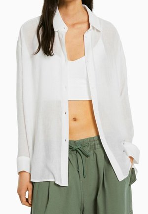 Vrouw draagt een doorschijnend wit overhemdbloesje over een witte cropped top en losse groene broek met trekkoord, staat met handen deels in zakken.
