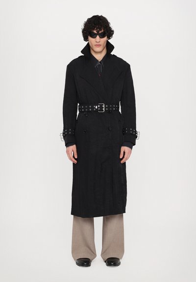 Schwarzer Trenchcoat mit doppelt geknöpften Knöpfen, breitem Kragen und Gürtel-Taille. Breite beige Hose und schwarze Schuhe mit glatter Oberfläche.