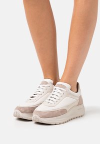 Person som bär beige och vita sneakers med lågt skaft, suède detaljer och tjocka gummisulor, stående mot en vit bakgrund.
