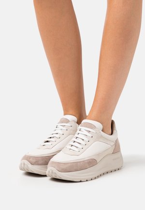 Persona que lleva zapatillas bajas en beige y blanco con detalles de ante y suelas de goma gruesa, de pie sobre un fondo blanco.