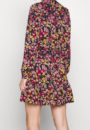 Femme portant une robe à fleurs à manches longues avec des fleurs roses, jaunes et rouges sur fond sombre, présentant un ourlet à volants et une taille ceinturée.