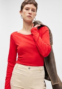 Piros, hosszú ujjú felső, széles nyakkivágással, világos bézs nadrággal párosítva. A felső puha, texturált felületű, és a modell egy barna dzsekit tart.