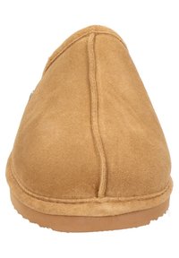 Beige suède pantoffel met een ronde neus, zachte textuur en een platte rubberen zool, met zichtbare stiksels langs de naad.