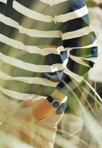 Inimene, kellel on seljas musta ja valget triibuline pikivarrukatega särk, käsi beežide pükste taskus, must kell randmel, udune muru esiplaanil.