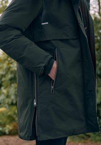 Manteau imperméable vert foncé avec des zippers sur les poches et une doublure texturée. Il présente un col haut et des sangles réglables sur les épaules.