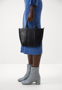 Black leather tote bag with dual handles, featuring vertical stitching. Model wearing a blue ribbed dress and light gray ankle boots with a block heel.