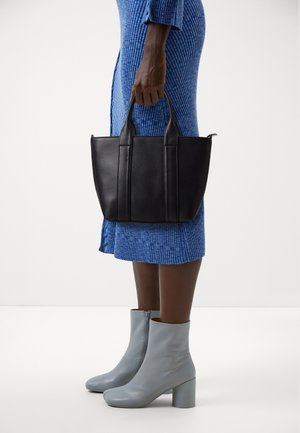 Black leather tote bag with dual handles, featuring vertical stitching. Model wearing a blue ribbed dress and light gray ankle boots with a block heel.