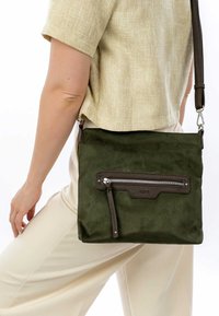 Person wearing beige top and pants carrying dark green suede crossbody bag with front zipper pocket and dark brown strap.