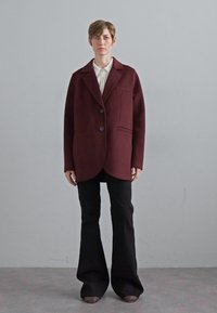 Blazer oversize bordeaux in misto lana con due bottoni e tasca frontale, abbinato a pantaloni neri a zampa d'elefante e scarpe basse a fantasia.