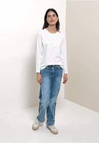 White long-sleeve shirt, light blue high-waisted jeans, and beige sneakers. Room with neutral-colored walls, smooth texture flooring.