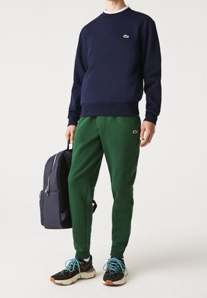 Personne portant un sweat-shirt marine et un pantalon de jogging vert avec des logos, tenant un sac à dos marine, debout en baskets avec des lacets bleus sur fond blanc.