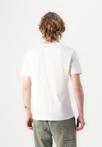 T-shirt bianco in cotone con maniche corte, collo tondo classico e una consistenza liscia. Manichino di spalle, abbinato a pantaloni grigi chiari.
