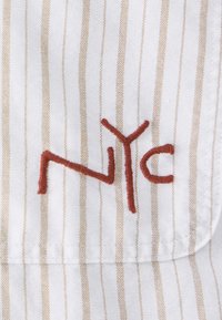 Tessuto bianco con strisce verticali marrone chiaro e la scritta ricamata "NYC" in filo marrone rossastro. Presenta una texture liscia.