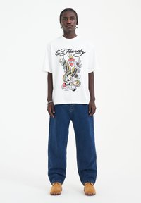Bílý tričko s barevným grafickým motivem Ed Hardy, který zahrnuje lebku a křídla, spárované s volnými modrými džíny a hnědou obuví.