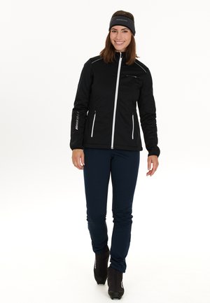 Veste noire à manches longues avec fermeture éclair blanche et accents réfléchissants, associée à un pantalon navy et des bottes noires. Inclut un bandeau noir.
